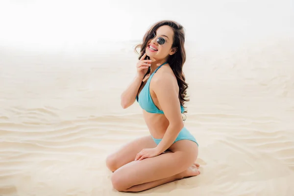 Beautiful smiling girl in blue Bikini and sunglasses sitting and posing on sandy beach — Stock Photo