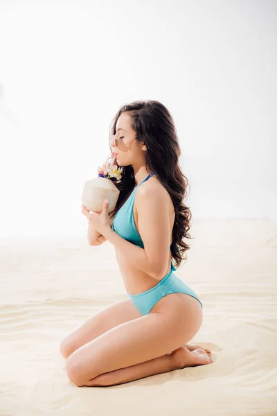 Hermosa chica sexy en bikini azul sentado y beber cóctel de coco en la playa de arena - foto de stock