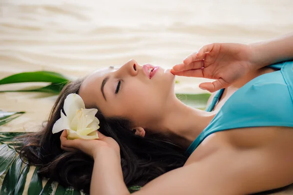 Schönes Mädchen mit geschlossenen Augen posiert mit Blume, während sie am Strand liegt — Stockfoto