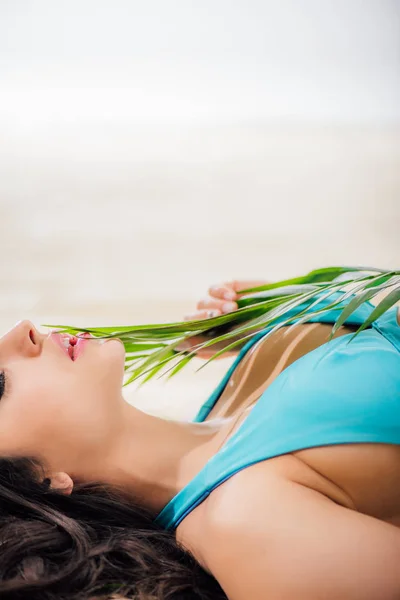 Cropped view of sexy girl in bikini with eyes closed posing with green leaf while lying on beach with copy space — Stock Photo