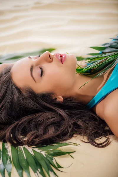 Красивая девушка с закрытыми глазами позирует с зеленым листом, лежа на пляже — стоковое фото