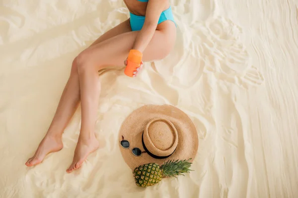 Vue du dessus de la fille en bikini avec crème solaire, chapeau de paille, ananas et lunettes de soleil se détendre sur la plage — Photo de stock