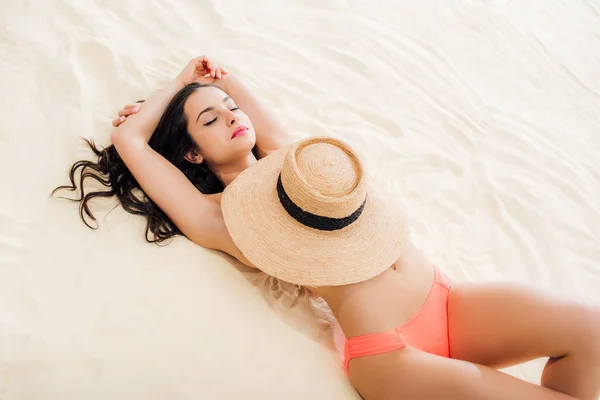 Sexy mujer joven con los ojos cerrados cubriendo los pechos con sombrero de paja mientras está acostado en la playa - foto de stock