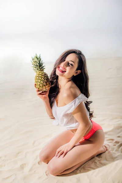 Hermosa chica sonriente sentado, mirando a la cámara y posando con piña en la playa de arena - foto de stock