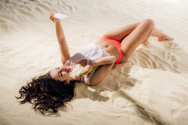 Красивая улыбающаяся девушка лежит на пляже с цветами и делает селфи на смартфоне — стоковое фото