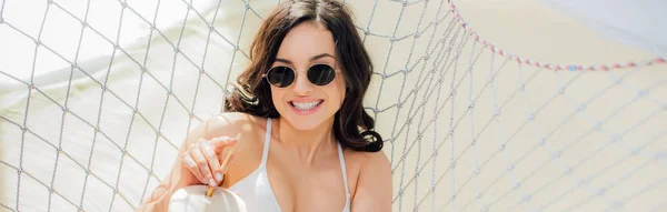 Panoramic shot of beautiful smiling girl in sunglasses with coconut cocktail lying in hammock on beach — Stock Photo