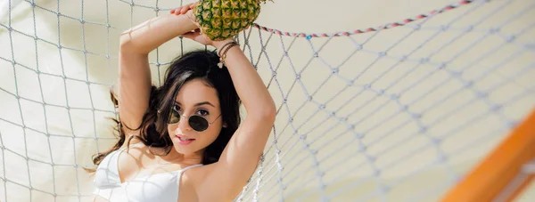 Panoramic shot of beautiful girl in sunglasses holding pineapple and lying in hammock on beach — Stock Photo