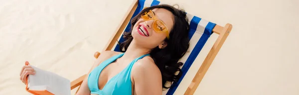 Plano panorámico de chica sonriente en bikini y gafas de sol celebración libro y relajarse en la tumbona en la playa - foto de stock