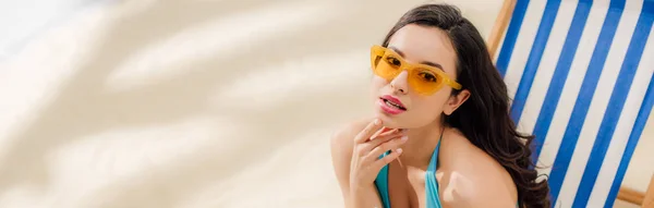Панорамный снимок девушки в бикини и солнцезащитных очках, отдыхающей на шезлонге на пляже с копировальным пространством — стоковое фото