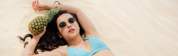 Plan panoramique de belle fille en bikini et lunettes de soleil posant avec de l'ananas tout en étant couché sur la plage — Photo de stock