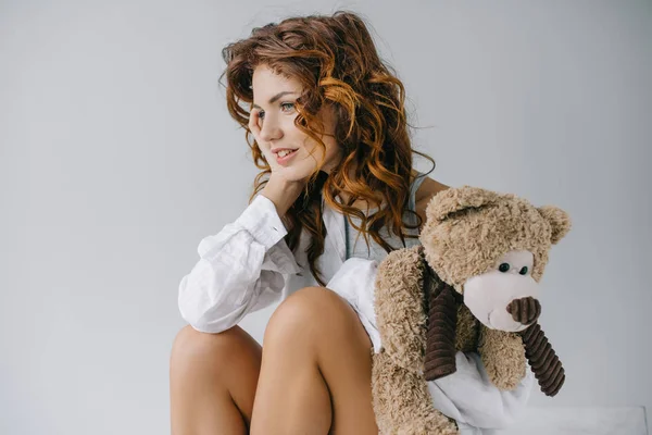Весела кучерява молода жінка тримає плюшевого ведмедя, сидячи на білому — стокове фото