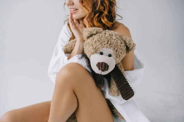Vue recadrée de joyeuse jeune femme bouclée tenant ours en peluche assis sur blanc — Photo de stock