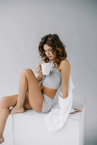 Chica con el pelo rojo rizado mirando a la taza de café mientras está sentado en gris — Stock Photo