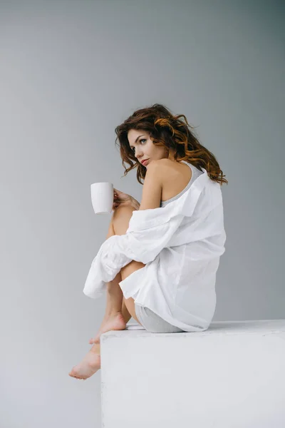 Atraente jovem segurando xícara com café e sentado em cinza — Fotografia de Stock