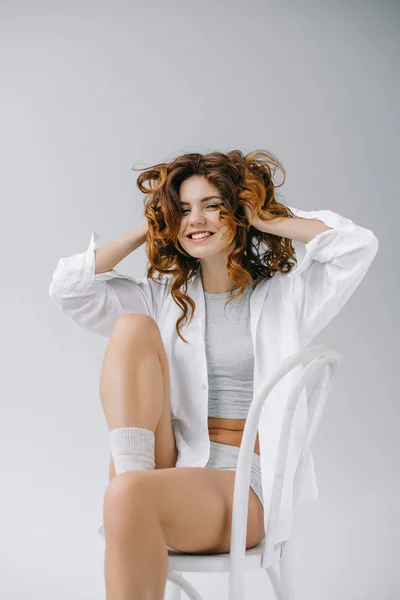 Cheerful girl touching red curly hair and smiling on grey — Stock Photo