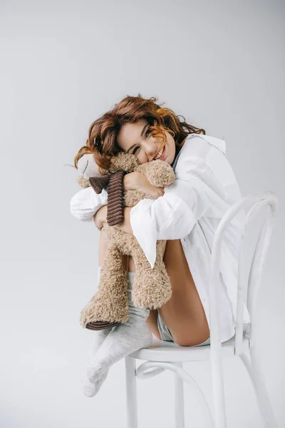 Glücklich lockige Frau hält Teddybär, während sie auf Stuhl sitzt — Stockfoto