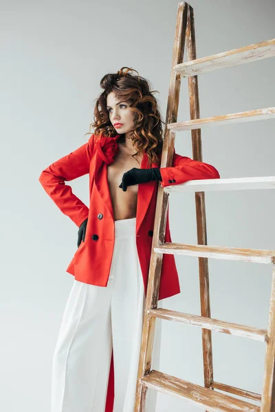 Tendance et belle rousse jeune femme debout avec la main sur la hanche près de l'échelle en bois sur gris — Photo de stock