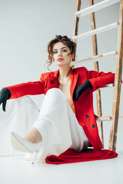 Enfoque selectivo de la mujer joven con estilo sentado cerca de escalera de madera y mirando a la cámara en blanco - foto de stock