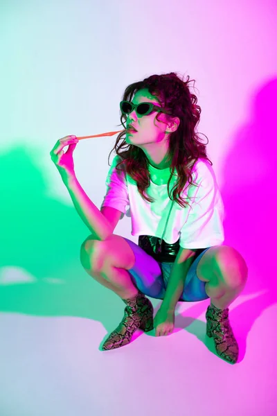 Attractive curly woman in sunglasses sitting with bubble gum on pink with illumination — Stock Photo