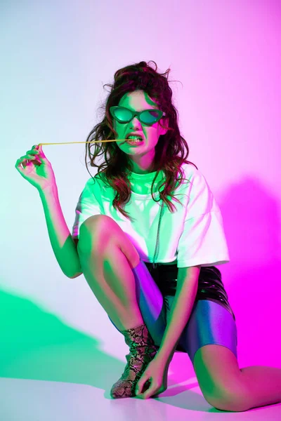 Curly young woman in sunglasses sitting with bubble gum on pink with illumination — Stock Photo