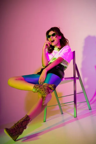 Mujer pelirroja elegante y feliz sentado en la silla y tocar gafas de sol en púrpura con gradiente - foto de stock