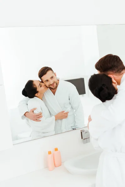 Selective focus of happy woman hugging and kissing cheek of man while looking at mirror — Stock Photo