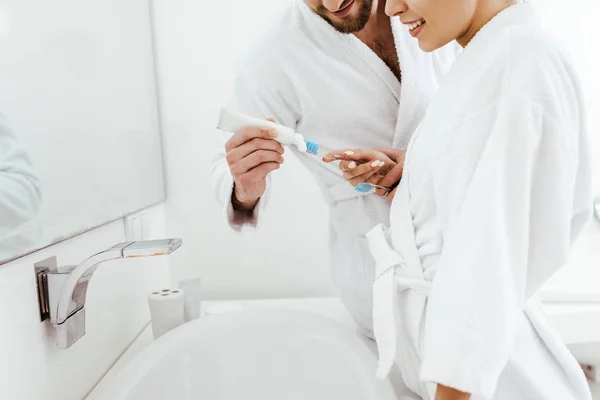Обрезанный вид бородатого мужчины, держащего зубную пасту рядом с веселой женщиной с зубной щеткой в руке — стоковое фото