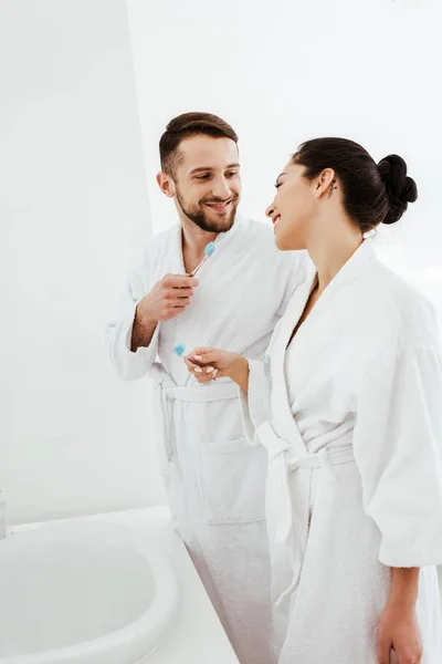 Happy man holding toothbrush and looking at brunette woman in bathroom — Stock Photo