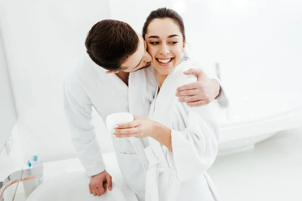 Alegre novio besos mejilla de feliz chica celebración contenedor con crema facial — Stock Photo