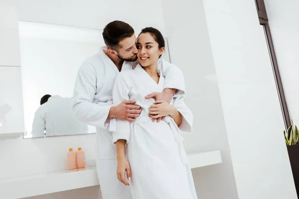 Handsome man kissing cheek of happy girlfriend standing in bathrobe — Stock Photo