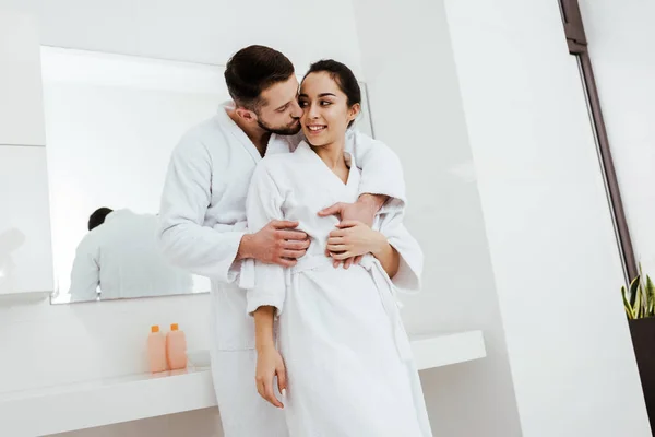 Hombre guapo besar mejilla de feliz novia de pie en albornoz — Stock Photo