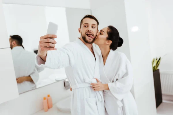 Girlfriend kissing cheek of cheerful man showing tongue while taking selfie in bathroom — Stock Photo