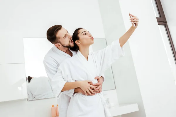Femme gaie avec visage de canard prenant selfie tout en se tenant avec petit ami dans la salle de bain — Photo de stock