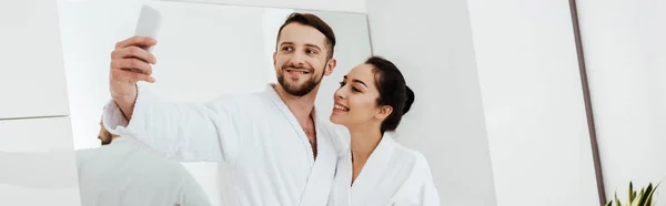 Panoramic shot of cheerful man taking selfie while standing with girlfriend in bathroom — Stock Photo