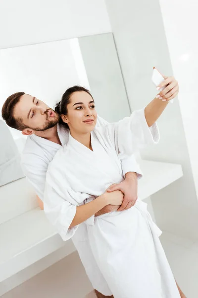 Alegre hombre con pato cara tomando selfie mientras abrazando novia en cuarto de baño - foto de stock