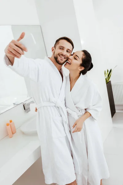 Alegre pareja tomando selfie y sonriendo mientras de pie en albornoces - foto de stock