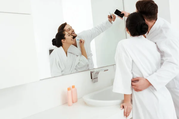 Hombre alegre abrazando a mujer feliz en albornoz mientras toma selfie en cuarto de baño — Stock Photo