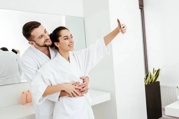 Alegre novio abrazando feliz novia tomando selfie en cuarto de baño — Stock Photo
