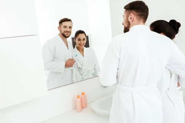 Messa a fuoco selettiva dell'uomo barbuto che tiene lo spazzolino da denti e guarda lo specchio con una donna bruna felice — Foto stock