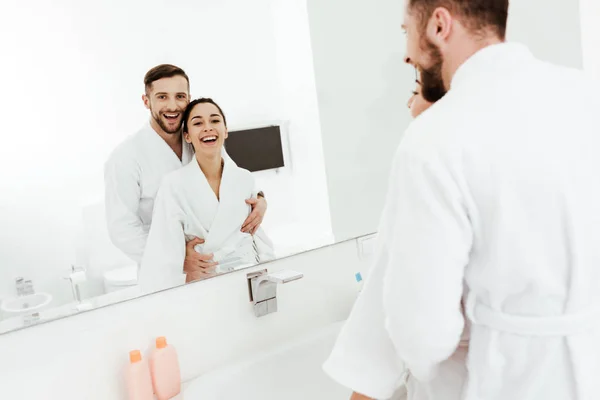 Selective focus of bearded man looking at mirror with cheerful brunette woman while hugging in bathroom — Stock Photo