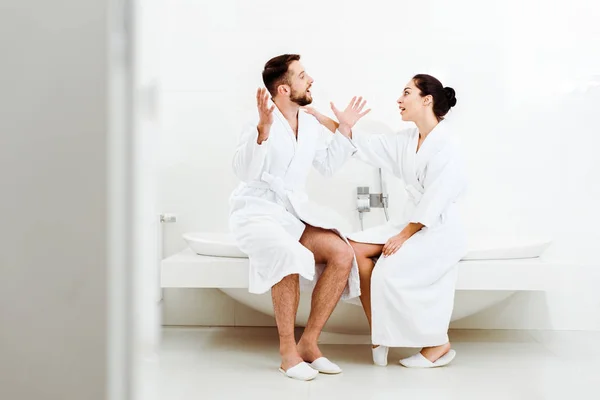 Foco seletivo de homem barbudo feliz sentado e gestual enquanto conversa com mulher atraente em roupão de banho — Fotografia de Stock