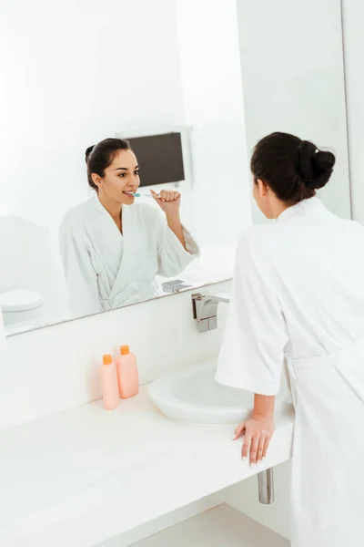 Messa a fuoco selettiva di felice giovane donna lavarsi i denti e guardando specchio in bagno — Foto stock