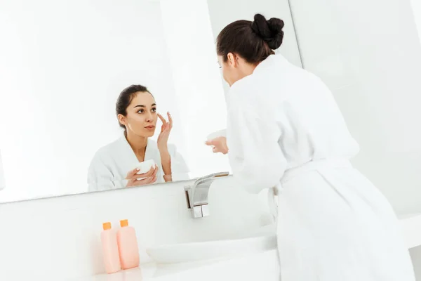 Selektiver Fokus der attraktiven brünetten Frau, die Gesichtscreme aufträgt, während sie in den Spiegel schaut — Stockfoto