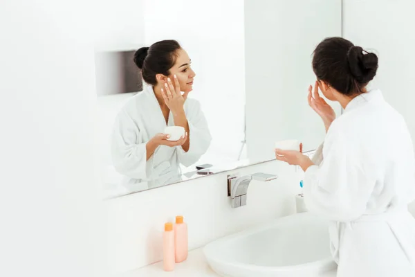 Fuoco selettivo della donna attraente che applica la crema per il viso mentre si guarda allo specchio — Foto stock