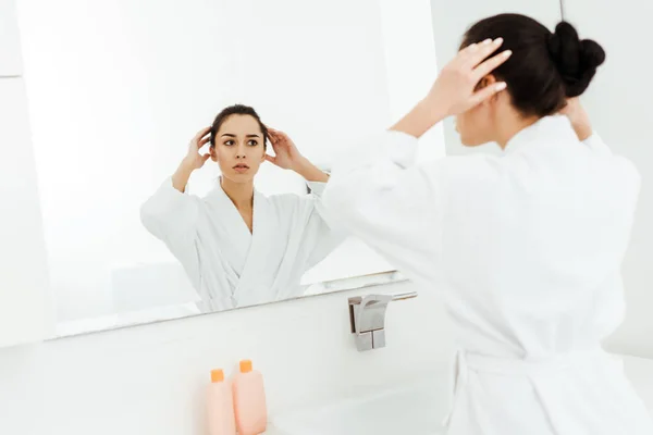 Selektiver Fokus der attraktiven brünetten Frau, die beim Anblick des Spiegels im Badezimmer Haare berührt — Stockfoto