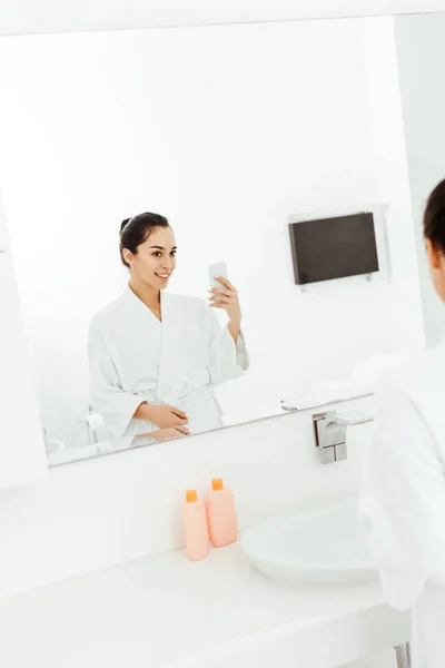 Селективное внимание привлекательной и счастливой молодой женщины, фотографирующей в ванной комнате — стоковое фото