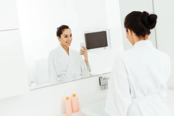Селективный фокус привлекательной и веселой молодой женщины, фотографирующей в ванной комнате — стоковое фото