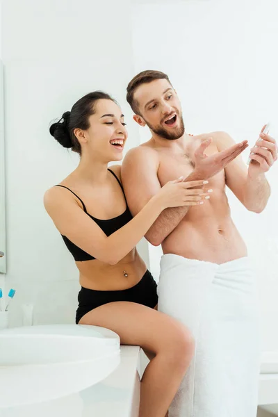 Mujer morena feliz en ropa interior mirando el teléfono inteligente en la mano de novio guapo - foto de stock