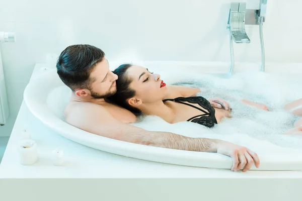 Hombre guapo acostado en la bañera con sexy mujer morena en sujetador de encaje - foto de stock