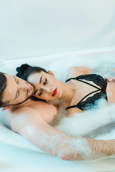 Bel homme torse nu couché dans la baignoire avec une jolie femme brune en soutien-gorge en dentelle — Photo de stock